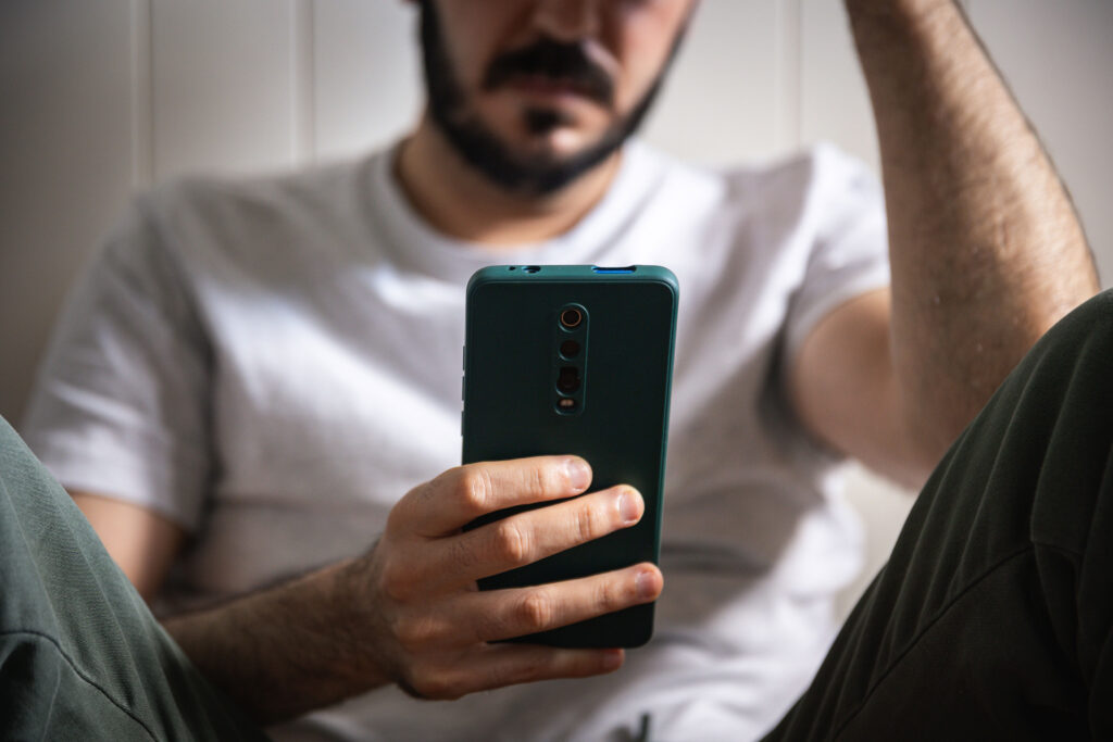 A man with a beard and wearing jeans and a t-shirt sits on the floor and looks at a phone in his hand.