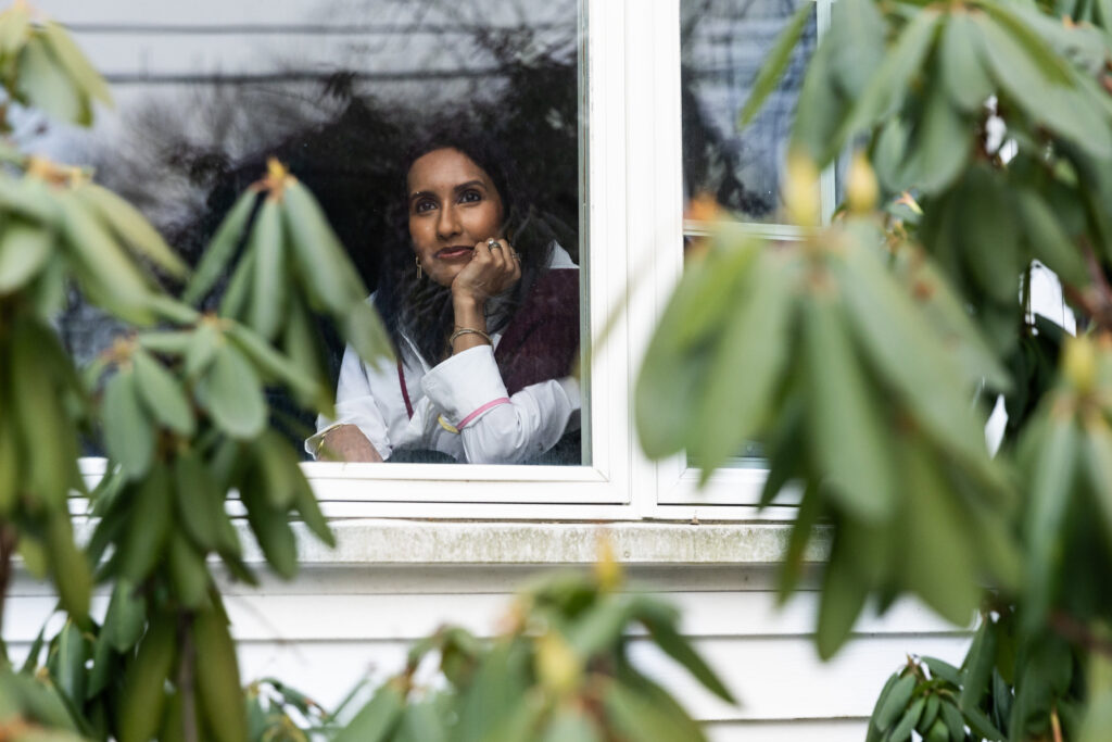 A woman looks out her window. Blurred greenery is seen in the foreground.