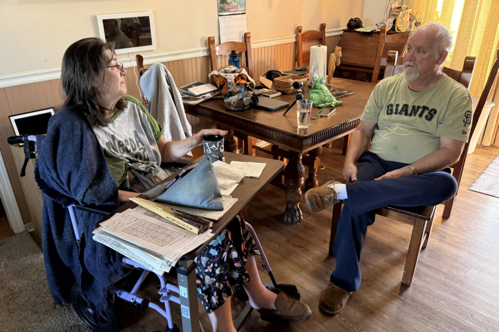 A husband sits in a chair conversing with his wife, who is in a wheelchair.