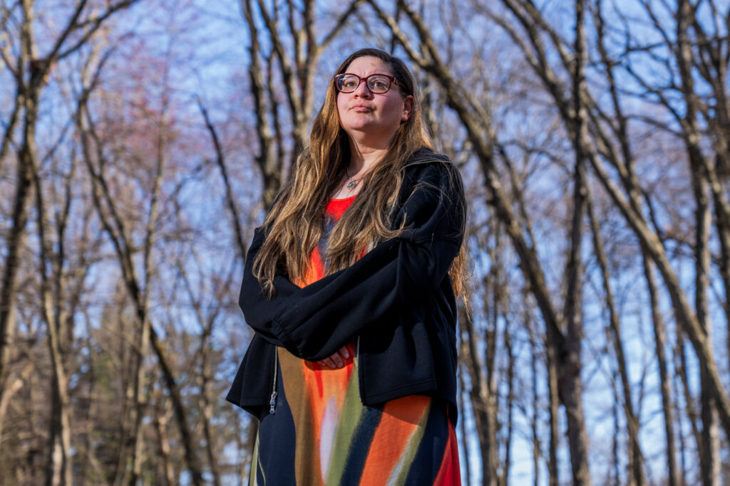 A woman stands outside in a wooded area.