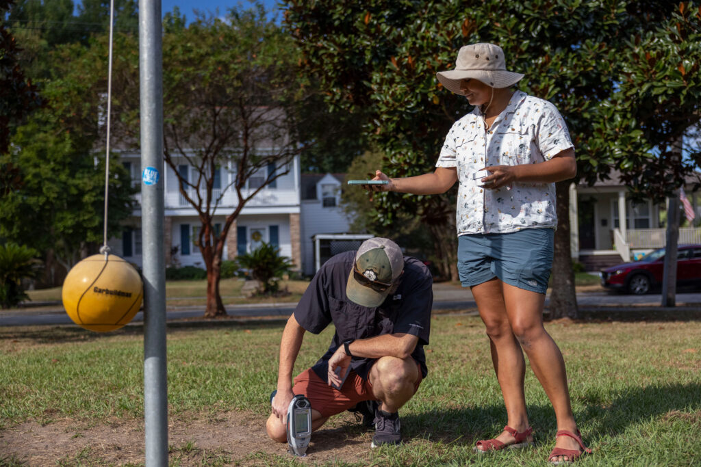 New Orleans Takes Steps To Assess and Clean Lead in Playgrounds After Investigation Two adults work outside. One on the left uses a device pressed to the ground to test for lead. Another holds out her arm, phone in hand, to get coordinates.