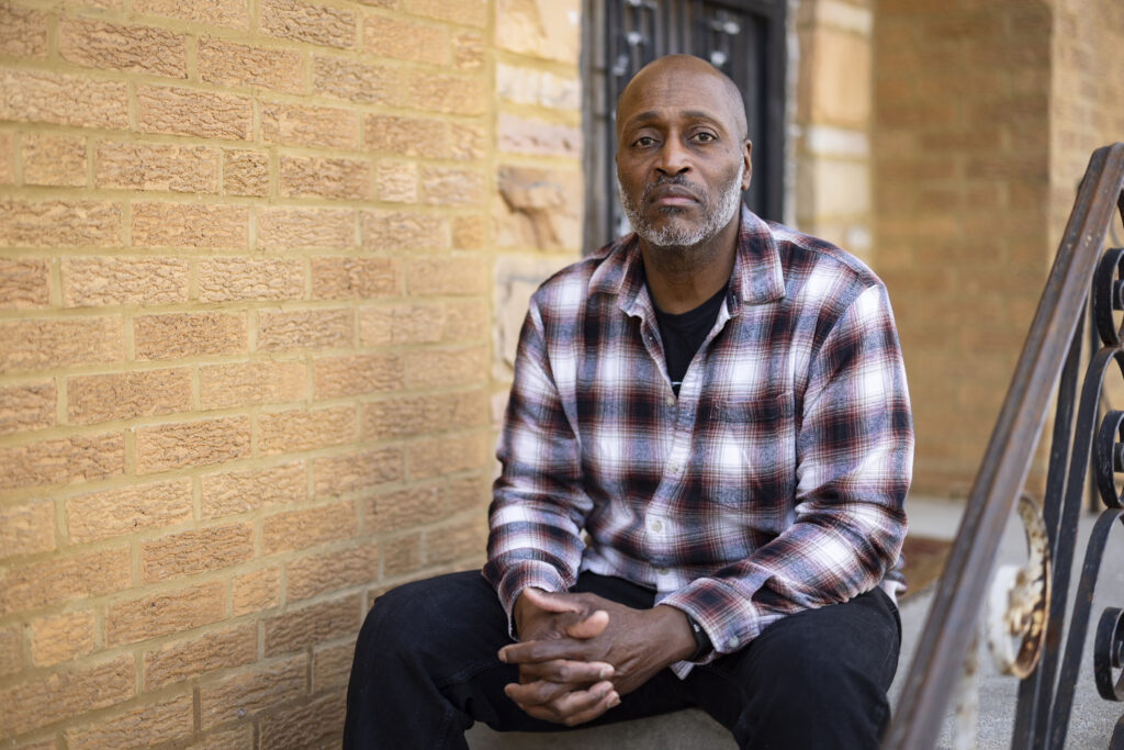 A man wearing a plaid button-up shirt sits on the front step of a home for a portrait