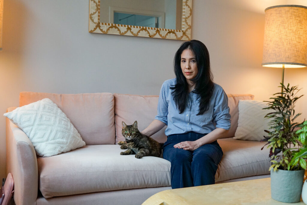 These Women Had Their Breasts Removed To Thwart Cancer. Then Came the Pain. A woman sits on a couch with a tabby cat beside her. Her hand gently rests beside her cat.